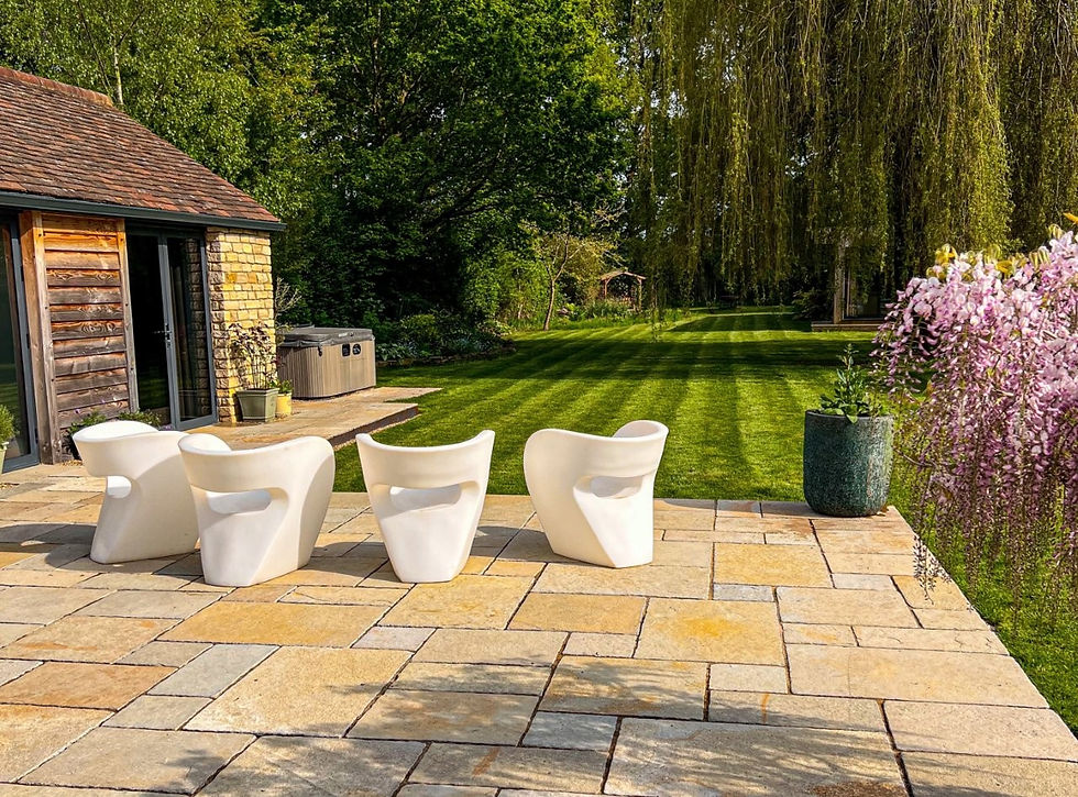 Patio with four white chairs on stone tiles, overlooking green lawn and trees. Rustic building on the left; pink flowers in foreground.