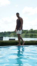 Man in a brown performance tank and cream shorts walking the edge of an infinity pool, lake behind him