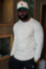 Man in a luxe living room wearing an Aesthetics Dynasty khaki Venture long sleeve, black Athletic Luxe joggers, and Dynasty Club cap, leaning confidently against furniture