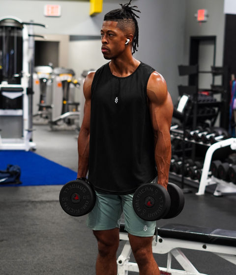 Man in gym holding dumbbells wearing Aesthetics Dynasty black athletic tank and green shorts, training