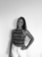 Jess, a Black British Caribbean woman, is stood against a plain wall with her hand on her hip. Her hair is dark and long, styled in goddess braids. She has a stripy halter neck strappy top on and white jeans and she's smiling to camera.
