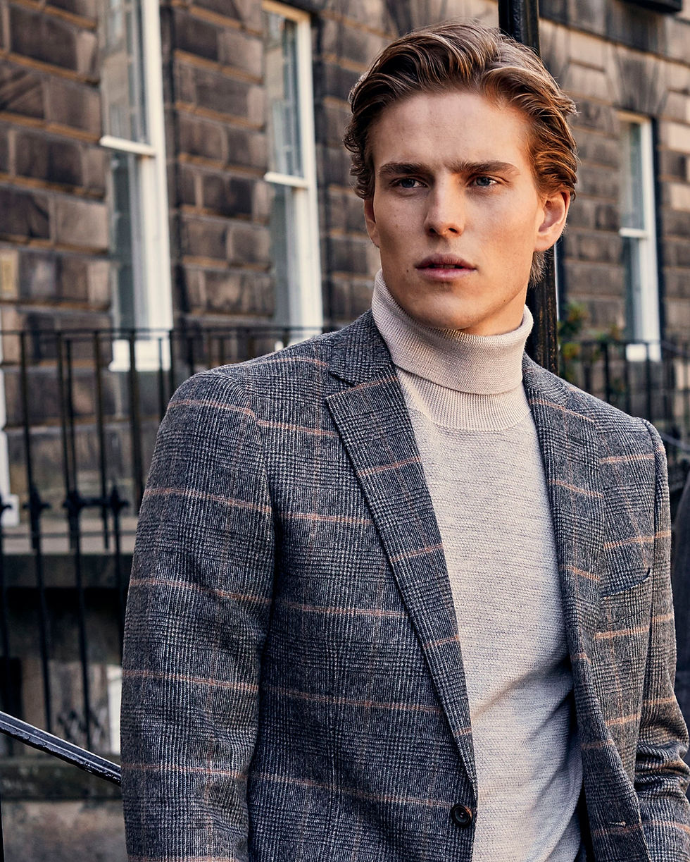 Ben is on a historic city street dressed in coordinated grey tailored clothing. Ben wears a checked blazer, He is a white male with dark blonde hair styled pushed back from his face.