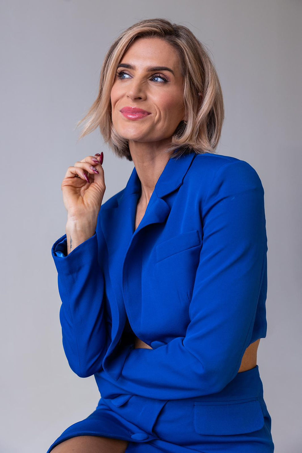 Sarah Jayne Dunn, a blonde female is standing against an orange backdrop. Her hair is up with small strands loose at the front and she is wearing a short sleeve white low cut dress with black lace trimming the bust. Her hands are in her pockets and she is looking towards the camera. 