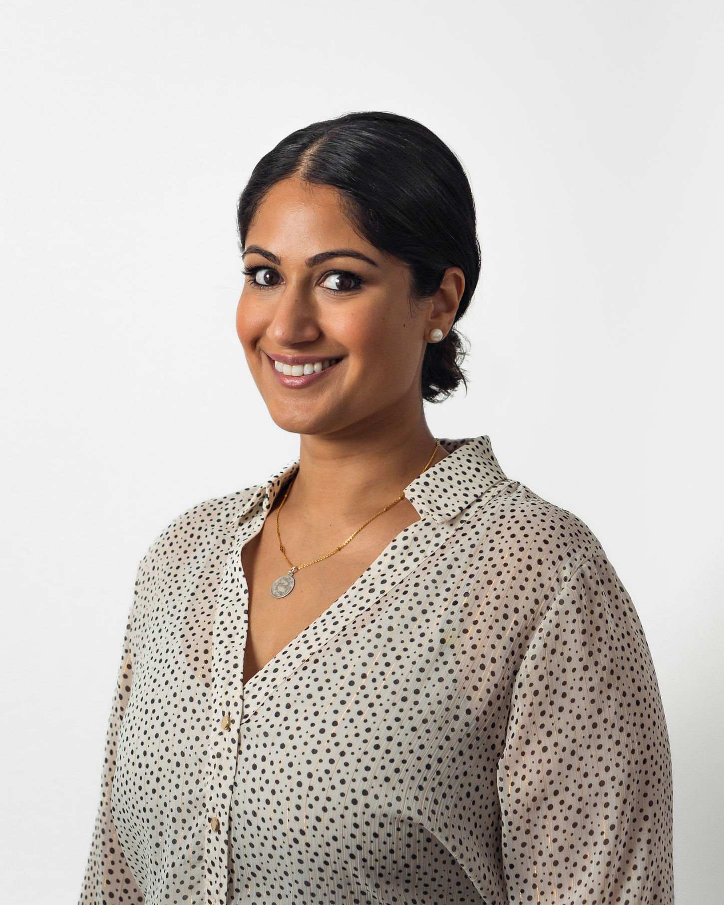 Amiee is a smiling woman with dark hair in a low bun, wearing a light-colored, polka dot blouse and a pendant necklace.