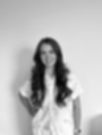 Ella Bamforth, a white female with long dark wavy hair. She is stood with her hand on her hip. She has a white blouse on with short sleeves and a faint squiggly pattern and she is smiling.