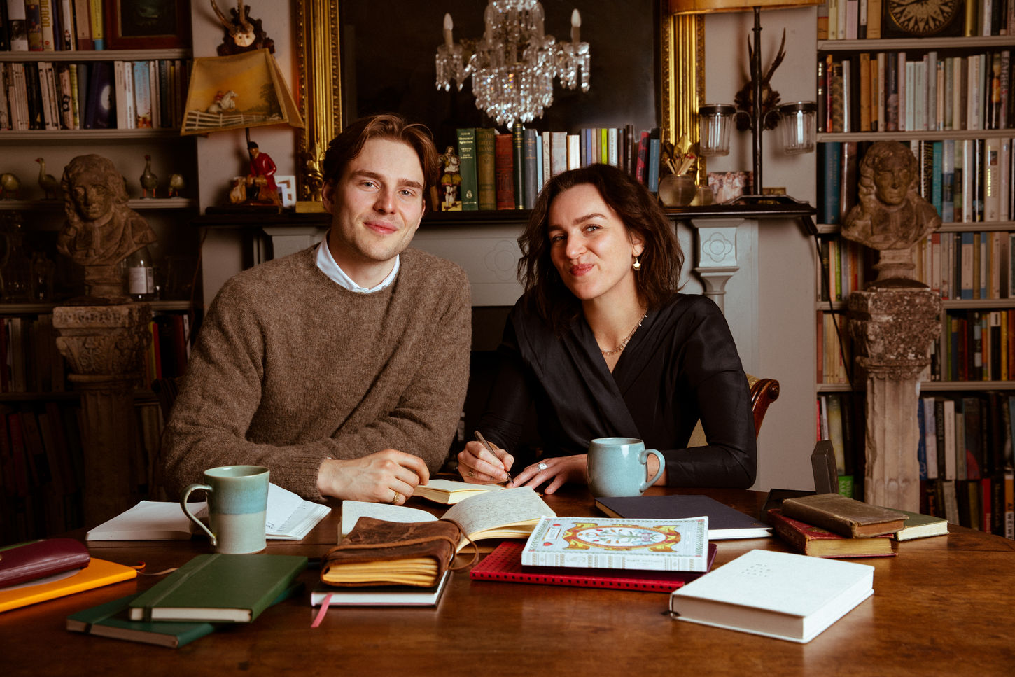 Ben and Mabel sit together at a wooden table in an elegant, book-filled library. They are writing in open notebooks, surrounded by books and ceramic mugs. The room features ornate furniture, classical sculptures, a chandelier, and richly decorated bookshelves.