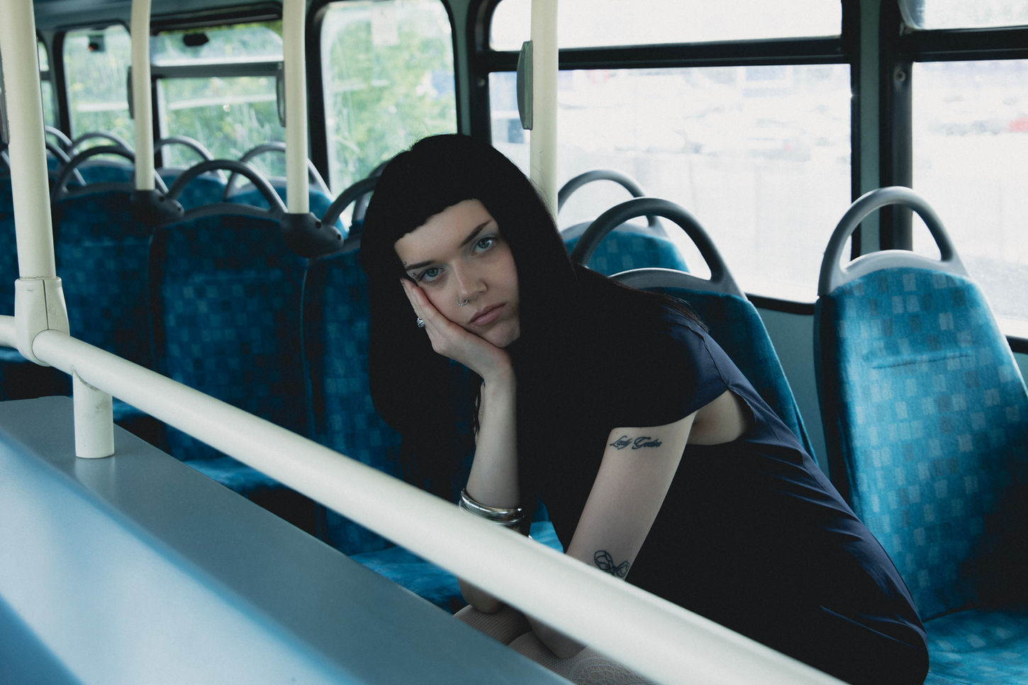 Zara is seen sitting on a blue TfL bus seat, behind railings. She has dark straight hair with micro bangs. Her head is tilted to the side with her head in her hand. She can be seen with multiple small tattoos on her arm. She is wearing thick silver bangles and rings.