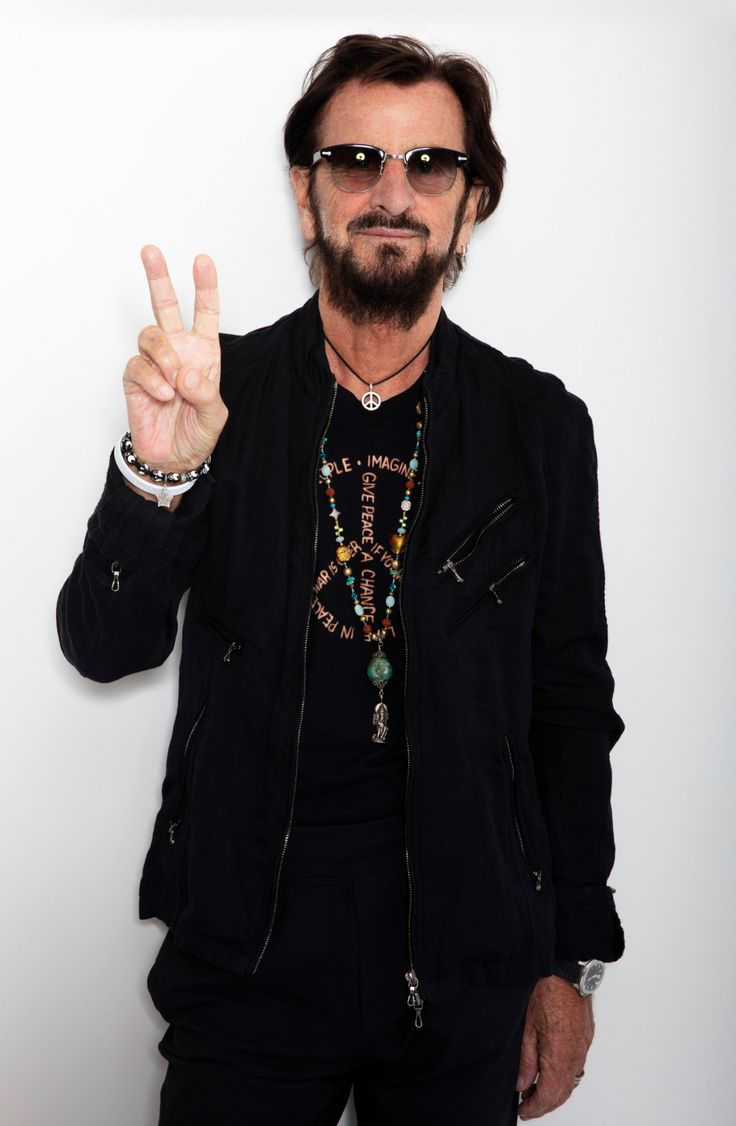 Ringo Starr in black jacket and sunglasses making peace sign. He's wearing colorful necklaces and a shirt with circular text. White background.