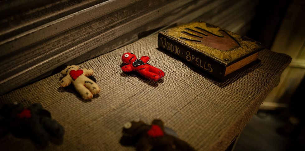 At Monster Quest Las Vegas. Voodoo dolls on burlap, one red with a bow tie, one beige with a heart. "Voodoo Spells" book nearby. Dimly lit, mysterious scene.