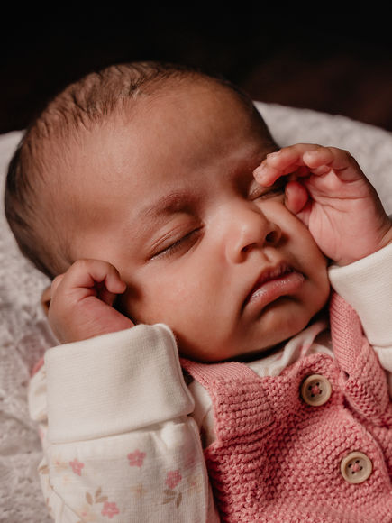 Schlafendes Baby in rosa Strickstrampler, liegt entspannt mit den Händen am Gesicht auf einer weißen Decke.