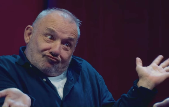 The British comedian, Bob Mortimer is seen pulling a face that indicated a feeling of "I don't know". He's sat, arms open, head tilted, wearing a blue shirt with a white t-shirt underneath. He's against a dark red background.