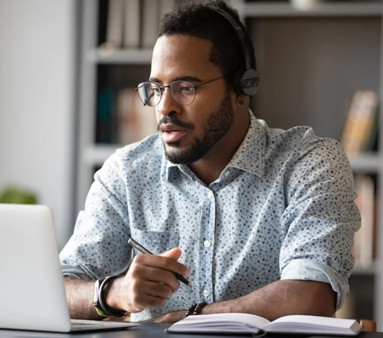 A young man of colour, is seen watching his laptop screen as he takes part in LMS neurodiversity training. He's wearing a smart, light coloured shirt and wire framed glasses.