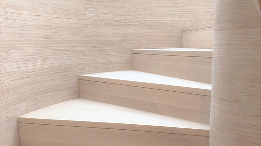 Modern trappa av ljust trä och plywood, med en mjuk, spiralformad gånglinje, placerad i en slät, rundad vägg med liknande struktur. Perfekt materialmatchning och mjuk geometri skapar en elegant, minimalistisk komposition.