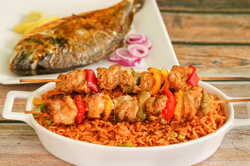 Skewers of grilled chicken and vegetables on a bed of jollof rice in a white dish; grilled fish and onion rings on a plate in the background.
