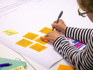 Women wearing glasses and black and white striped T-shirt writing on an orange post-it note on a large sheet of white paper with multiple post-its