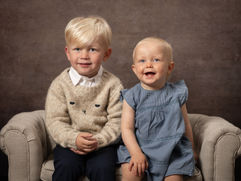 Två småsyskon sitter glada i en liten barnsoffa. Barnfotografering, syskonfotografering. Fotograf Camilla Geidemark Köping