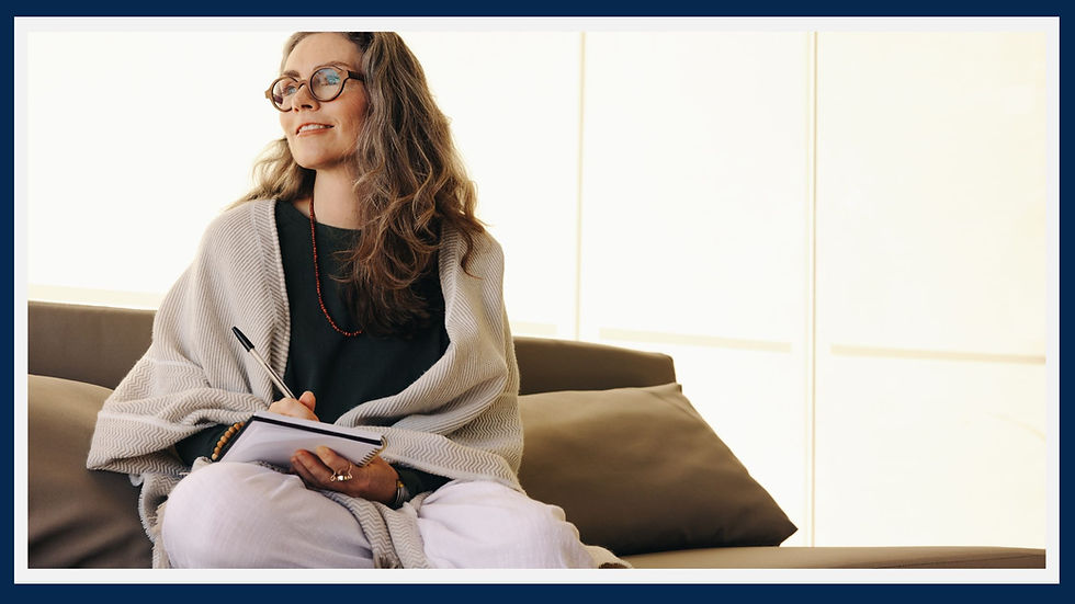 A woman sitting on a coach wrapped in a blanked and writing in her journal. She looks happy and confident.