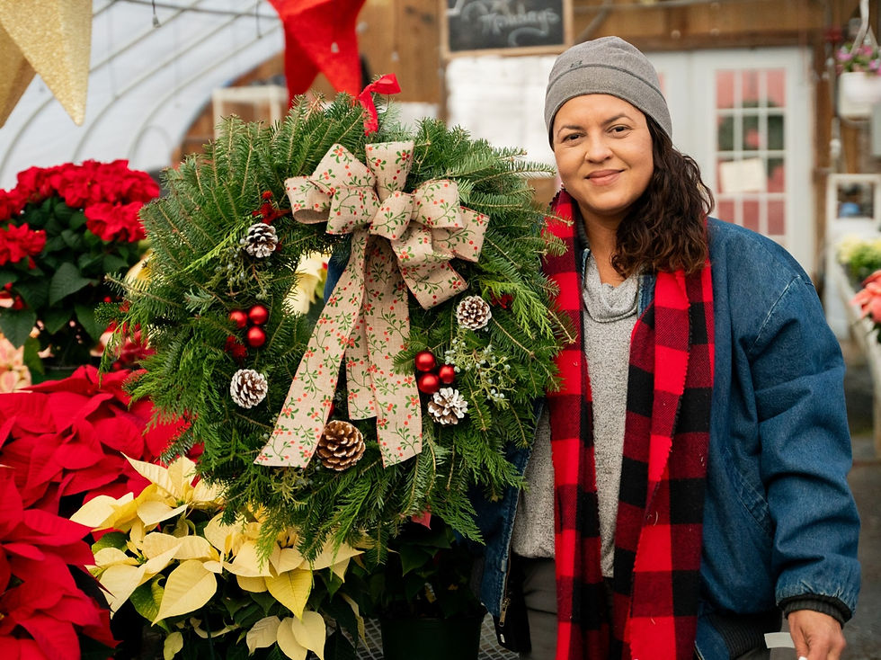 Woman holding large decorated balsam Christmas wreath in greenhouse surrounded by poinsettias and other Christmas plants