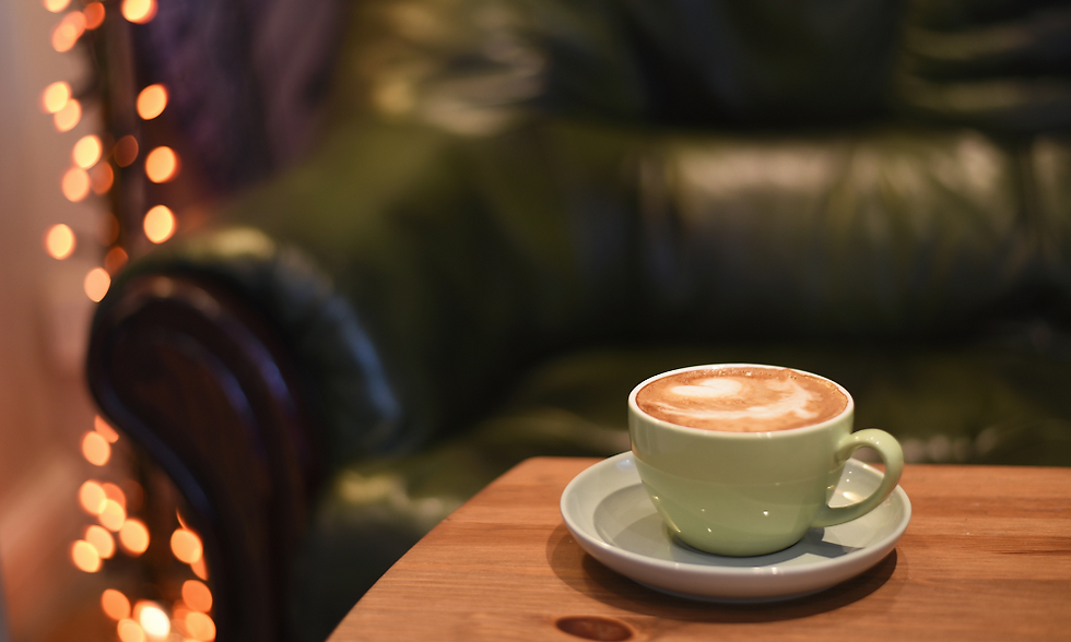 A Chelsea Coffee latte in a classic green mug sits on a wooden table with a plsuh leather chair and string lights in the background.