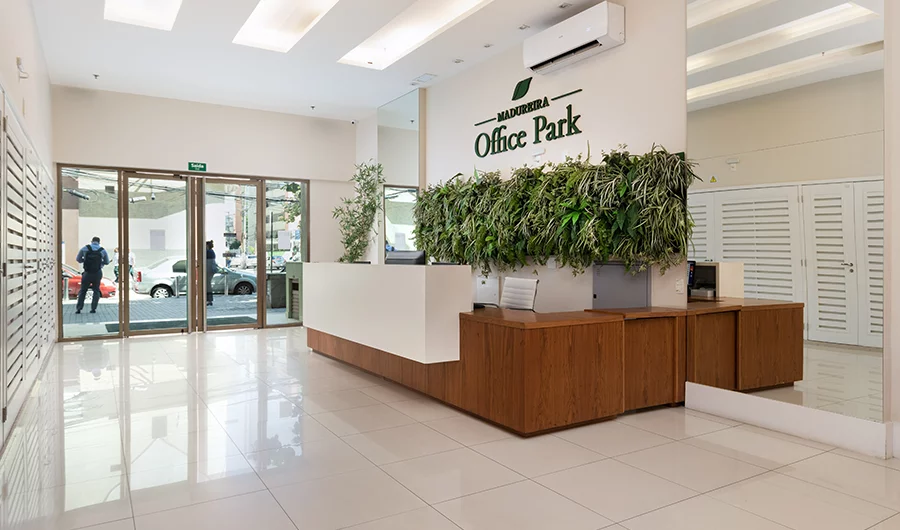 Recepção moderna com balcão em madeira e painel de plantas verdes, piso claro e portas de vidro que dão acesso à rua. Letreiro 'Office Park' na parede atrás do balcão.