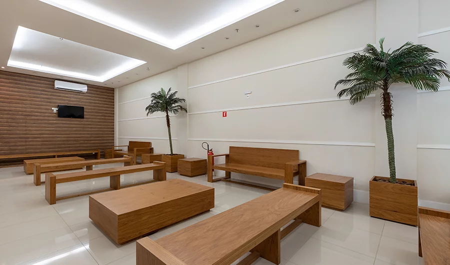 Sala de espera ampla com bancos e mesas de madeira, plantas decorativas em vasos altos, paredes brancas com molduras e iluminação embutida no teto.