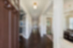 Elegant empty foyer with dark wood floors, white columns, open French door, and brass chandeliers leading to a bright hallway