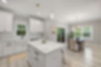 Bright kitchen with white cabinets, island, and pendant lights. Dining area with wooden table, gray chairs, and large window view of greenery.