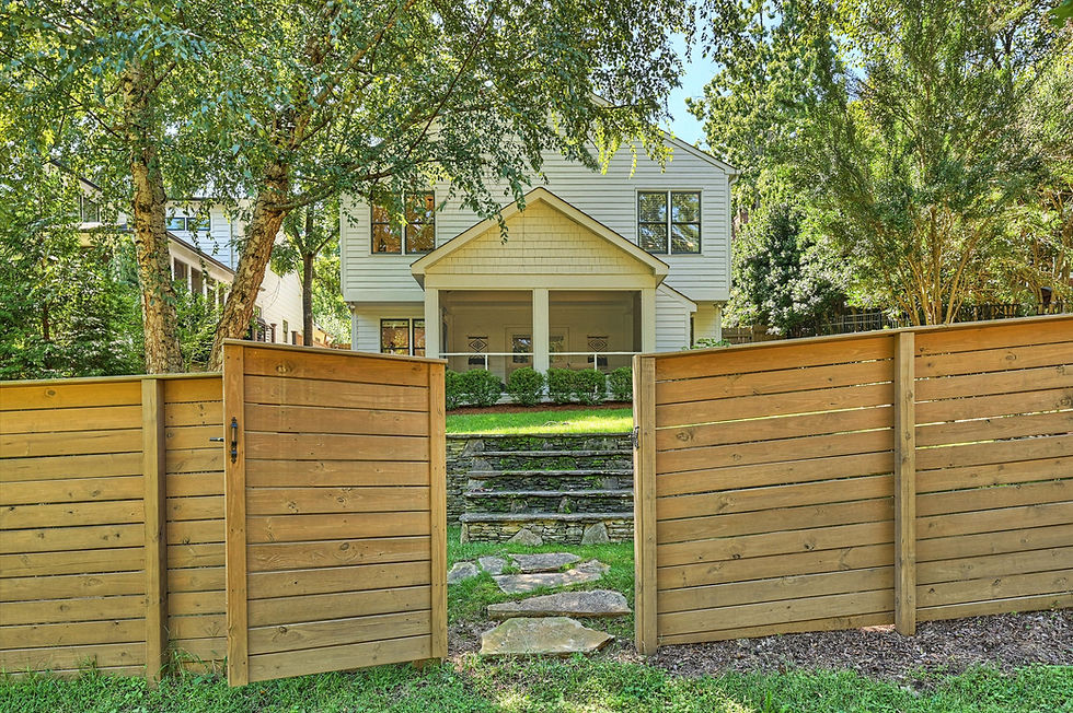 Open wooden gate frames a white two-story house with porch, stone steps, and lush trees in a sunny yard.
