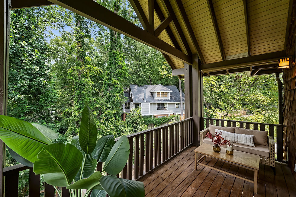 Covered wooden porch with beige sofa and plants overlooks lush trees and a gray house on a sunny day