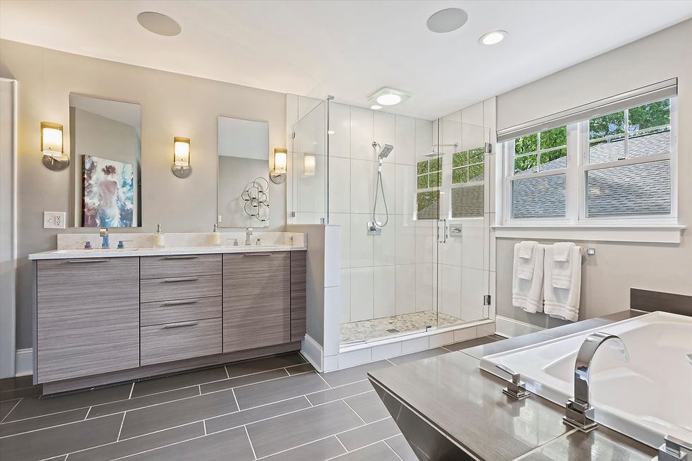 Modern bathroom with grey cabinets, twin sinks, glass-enclosed shower, large bathtub, and grey tile floor. Bright, clean, and spacious.