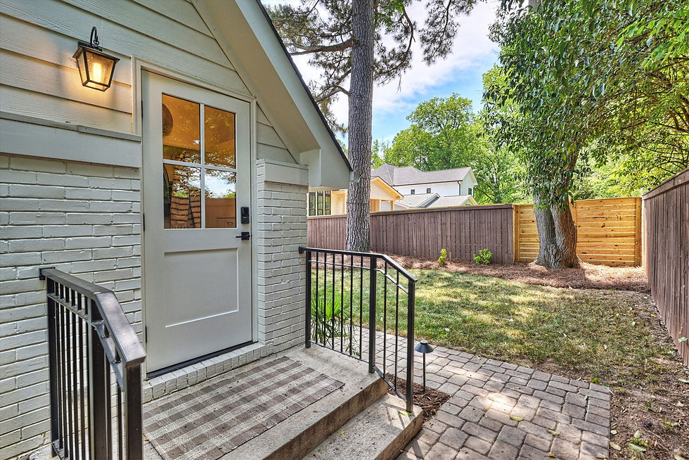 Cozy backyard with a white brick house, lantern-lit door, and metal railing. Wooden fence, trees, and sunny sky in the background.