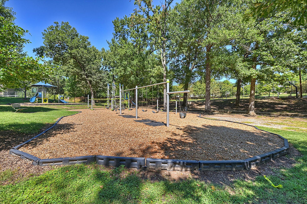 Empty park playground with swings, slides, and wood chips under tall trees on a sunny blue-sky day.