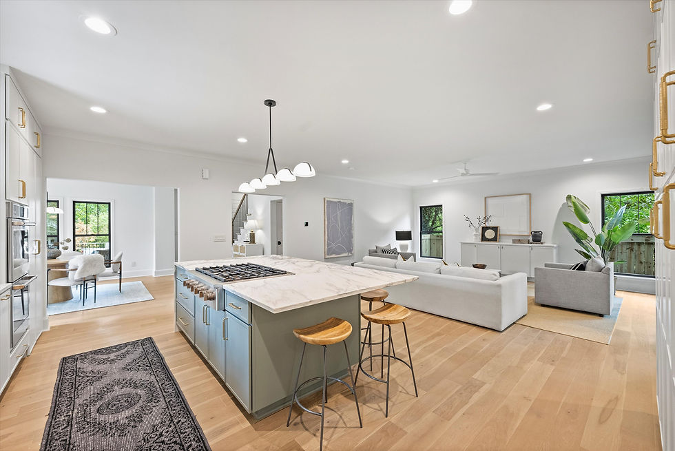 Modern kitchen and living room with wooden floors, marble island, stools, gray sofa, large windows, and green plants. Bright and airy ambiance.