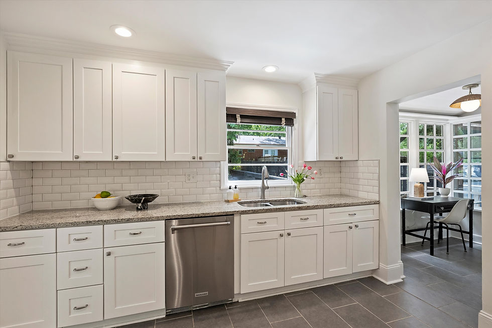 Modern kitchen with white cabinets, marble countertops, and stainless steel dishwasher. Window view and adjacent room with desk and lamp.