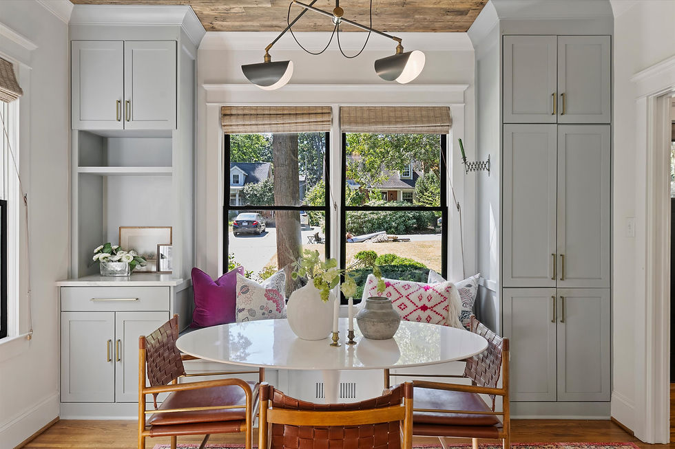 Bright built-in breakfast nook with round white table, woven chairs, colorful pillows, and large windows overlooking a leafy street.