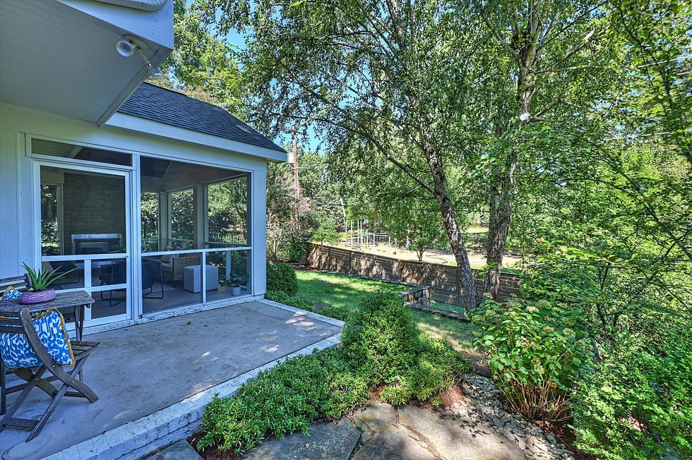 Sunny backyard patio beside a screened porch, with blue patterned chair cushions, lush green trees, and a quiet residential view.