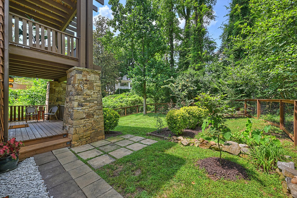 Wooded backyard with stone patio under a deck, two chairs, lush green lawn, trees, and fenced garden in bright daylight.