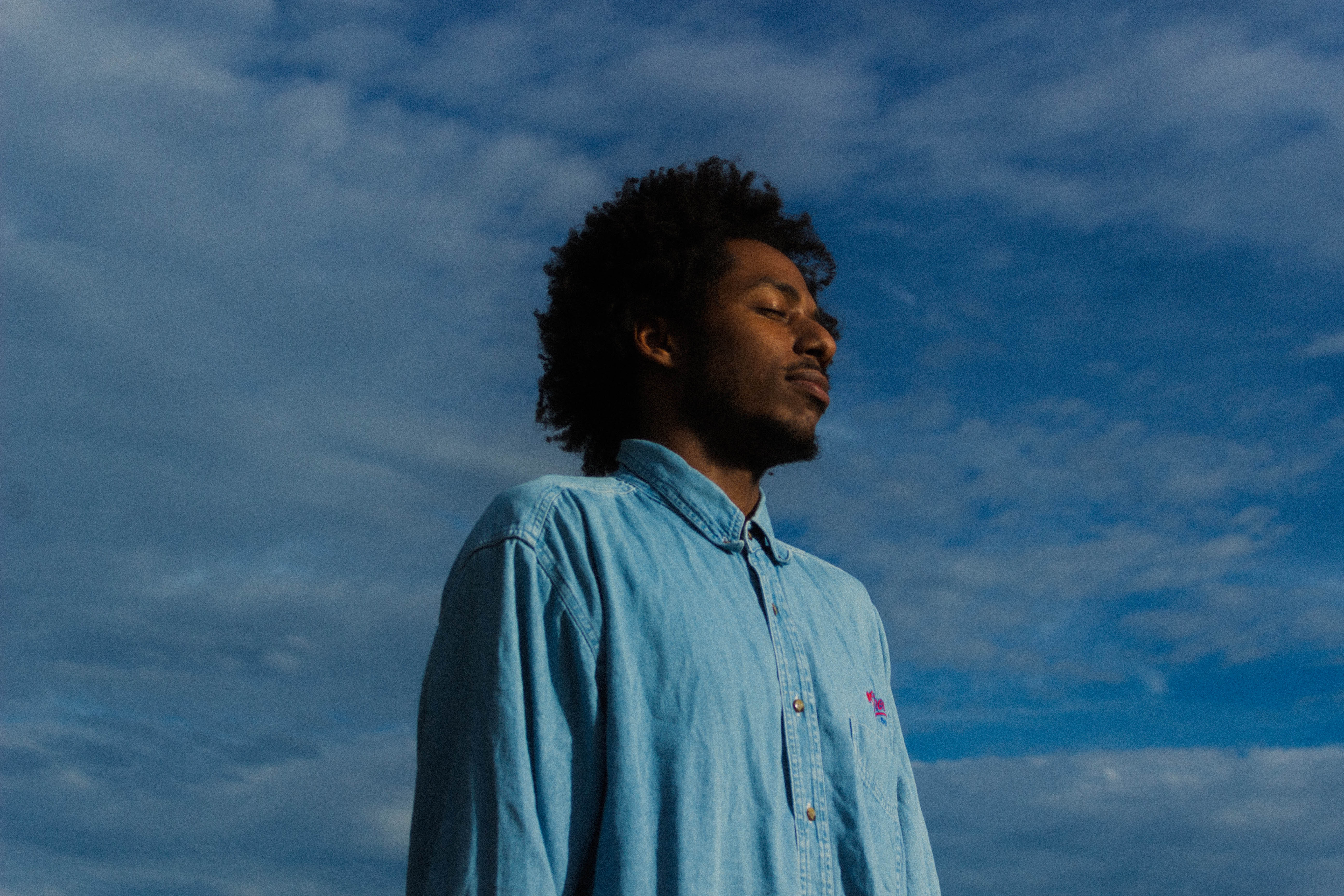 Person standing outdoors with eyes closed, wearing a light blue denim shirt, against a backdrop of a blue sky.