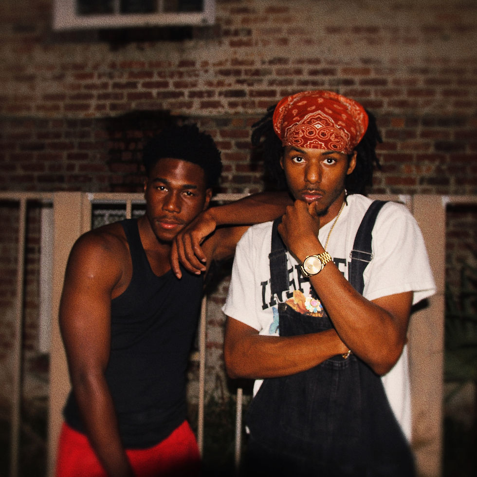 Two young men posing outside in front of a brick wall, one in a black tank top and red shorts, the other in a white T-shirt