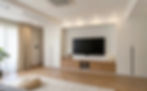 Modern living room with a wall-mounted TV, white cabinets, wooden floor, beige curtains, a large plant, and a woven pouf on a rug.