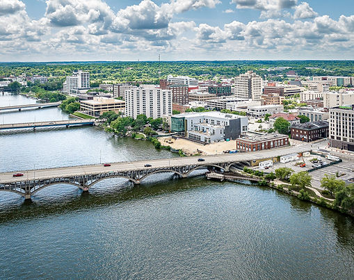 A wide-angle aerial shot of Rockford, Illinois, looking over the Rock River. The water is a dark, reflective blue-green, crossed by three distinct bridges that lead into a skyline of brick and modern office buildings. The background stretches into a vast green horizon under a dynamic sky with white puffy clouds. The image conveys a sense of a peaceful but active mid-sized American city called Rockford, IL.