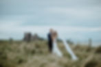 A bride and groom smiling in fron of Tantallon Castle near Edinburgh. They are freshly married. They got eloped in the countryside near Seacliff Beach with a view to Tantallon Castle. The bride is wearing a long veil which is flowing in the wind and a white simply beautiful wedding dress.