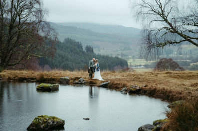 Bride and groom embracing by a peaceful Highland loch near Glen Affric, Scotland, during a cosy winter wedding, with misty mountains, forested hills and soft overcast skies creating a romantic Scottish Highlands backdrop.