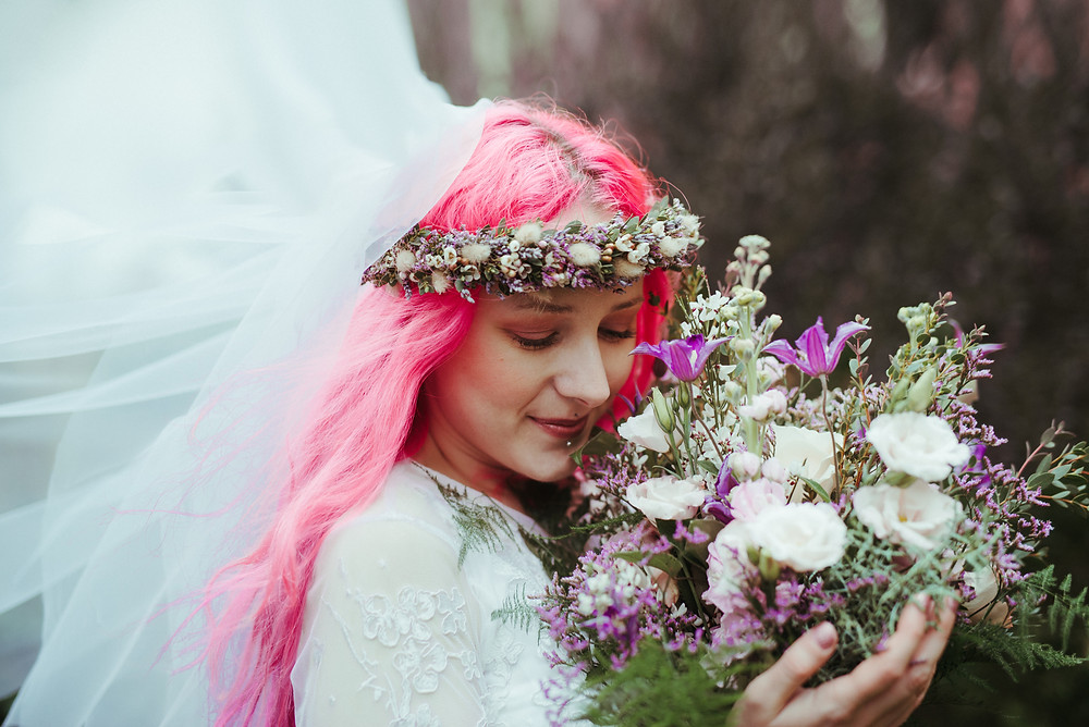 An alternative bride with a flowers crown and pink hair in Edinburgh at Holyrood Park. She is wearing a white flowing wedding dress with lace sleeves is from ASOS. Her wedding flower crown and the wedding bouquet is from Snapdragon Edinburgh.