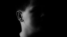 Silhouette of a person in deep shadow with a dark background. Only the left side of the face and neck are slightly illuminated.