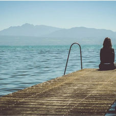 A person sits alone on a wooden dock, overlooking a calm lake and distant mountains under a clear sky, creating a serene mood.