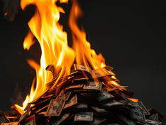 Pile of U.S. dollar bills burning with bright orange flames against a dark background, evoking waste and urgency.