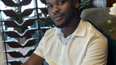 Smiling man in a white shirt sits in a room with wooden decor and plants. Soft lighting and a relaxed mood are evident.