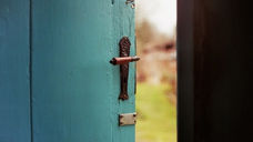 A blue door is slightly ajar, revealing blurry green and brown scenery outside. The mood is serene and inviting, with vintage hardware.