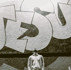 man looking up to the sky in front of a wall with the name Jesus painted graffiti style on it.
